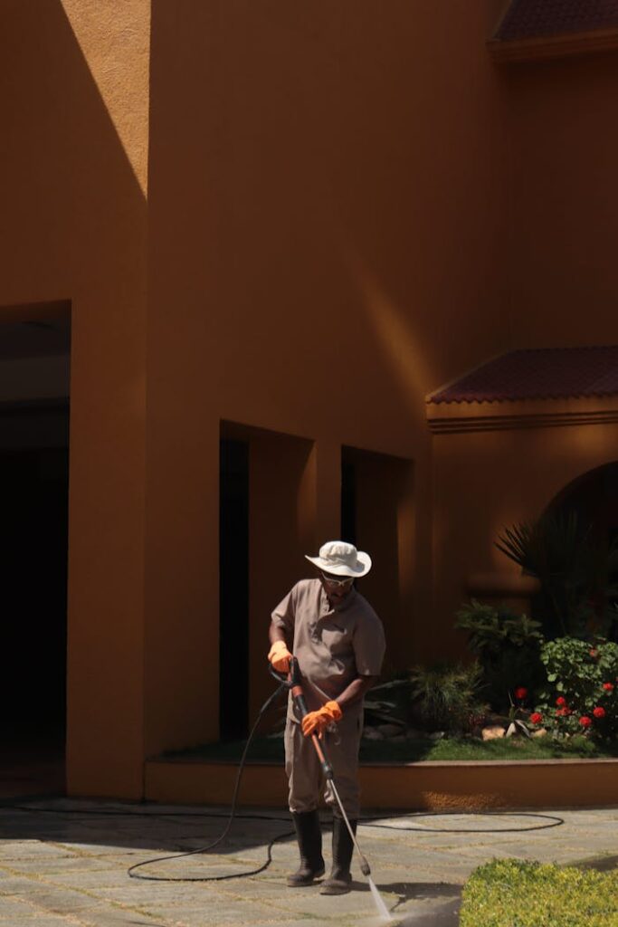 A gardener wearing a hat cleans an urban sidewalk with a pressure washer.